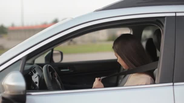 Egy fiatal nő ül egy kocsiban és biztonsági övet visel. Mosolyog és a kamerába néz. A kormányon tartja a kezét. 4K — Stock videók