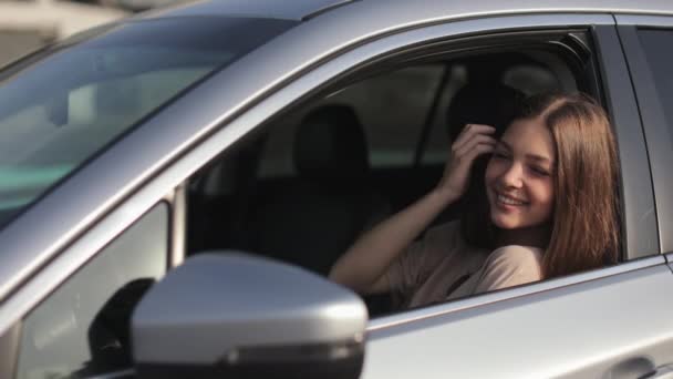 Una giovane donna è seduta in macchina sul sedile del conducente. Si guarda allo specchio laterale e si aggiusta i capelli con la mano. Sta sorridendo. 4K — Video Stock
