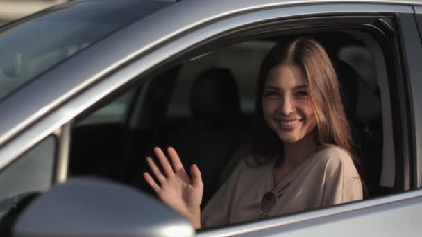 Una joven está sentada en el coche en el asiento del conductor. La mujer sonriente está mirando a la cámara y agitando su mano. El sol brilla sobre ella. 4K — Vídeos de Stock