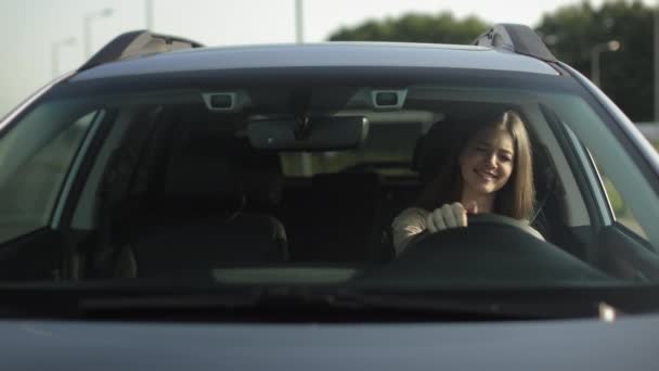 Una mujer feliz está sentada en su coche en el asiento del conductor. Está escuchando música y girando el volante. Ella sonríe. 4K — Vídeos de Stock