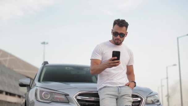 Otoparkta genç bir adam arabasının yanında duruyor. Akıllı telefonuyla sosyal ağlarda mesajlaşıyormuş. Güneş gözlüğü takıyor. 4K — Stok video