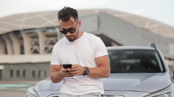 한 젊은 남자 가 주차장에 있는 자기 차 근처에서 있습니다. 그는 스마트폰으로 소셜 네트워크에 문자를 보내며 웃고 있습니다. 머리를 들고 카메라를 보고 있습니다. 4K — 비디오