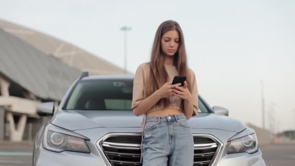 한 젊은 여자 가 주차장에 있는 자기 차 근처에서 있습니다. 그녀는 스마트폰으로 소셜 네트워크에 문자를 보내고 있습니다. 카메라가 그녀 주변을 움직이고 있습니다. 4K — 비디오