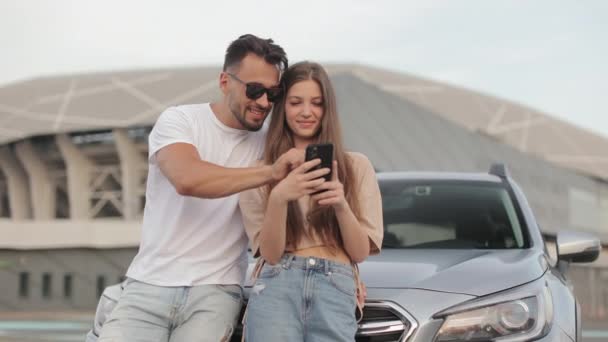 Bir adam ve bir kız bir arabanın yanında duruyorlar. Gülümsüyorlar ve akıllı telefonlarından bir şey izliyorlar. Kamera onların etrafında hareket ediyor. 4K — Stok video