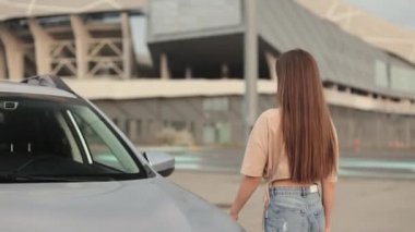 Genç bir kadın arabaya gidiyor. O, kapıyı açıyor ve sürücü koltuğunda oturuyor. Otoparktaki araba. Kamera onun yanına doğru hareket ediyor. 4K 50fps