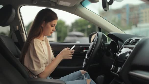 A young woman is sitting in the drivers seat. She is texting on her smartphone. Shooting from the side seats. 4K 50fps — Stockvideo