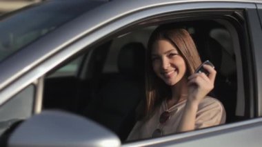 Başarılı bir kadın sürücü koltuğunda arabasında oturuyor. Arabasının anahtarını gösteriyor ve gülümsüyor. O, kameraya bakıyor. 4K