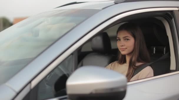 A young woman is sitting in the car and smiling. She is turning her head and looking at the camera. She is keeping her hands on the steering wheel. The camera is moving around. 4K — Wideo stockowe