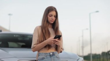 Otoparkta genç bir kadın arabasının yanında duruyor. Akıllı telefonuyla sosyal ağlarda mesajlaşıyor ve kafasını kaldırıyor. Etrafına bakıyor ve gülümsüyor. 4K