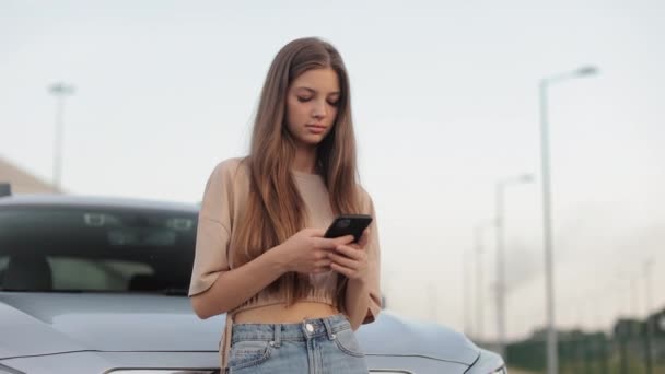 Otoparkta genç bir kadın arabasının yanında duruyor. Akıllı telefonuyla sosyal ağlarda mesajlaşıyor ve kafasını kaldırıyor. Etrafına bakıyor ve gülümsüyor. 4K — Stok video