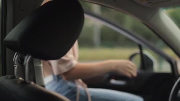 A young woman is sitting in a car in the drivers seat. She is taking off her mask and starting the car. Shooting from the back seats. 4K 50fps — Vídeo de stock