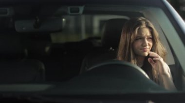 A tired woman is sitting in her car in the drivers seat. She is waiting for someone and leaning against the door. The sun is shining on her. 4K