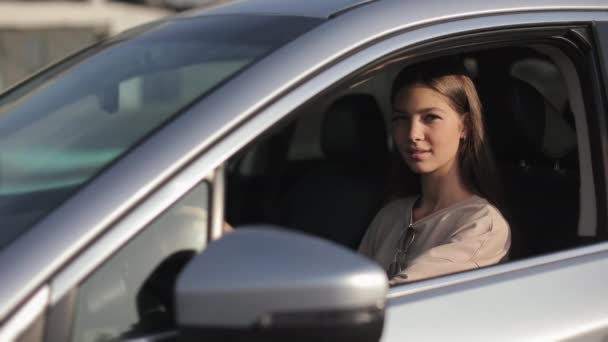 A young woman is sitting in the car in the drivers seat. She is turning her head and laughing. She is keeping her hands on the steering wheel. She is looking at the camera through the open window. 4K — Wideo stockowe