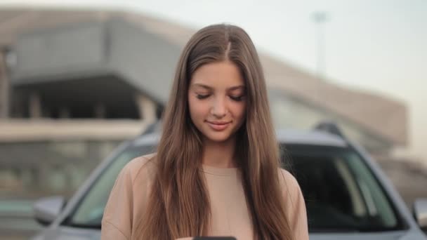 Portrait of a young woman who is standing near her car in the parking lot. She is texting on social networks. She is looking up at the camera with a smile. The camera is zooming in. 4K – stockvideo
