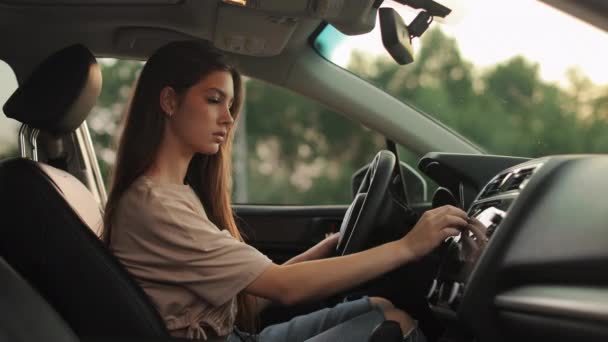 A young woman is sitting in the drivers seat. She is selecting music in the cars multimedia. She is smiling. Shooting from the side seats. 4K 50fps — Wideo stockowe