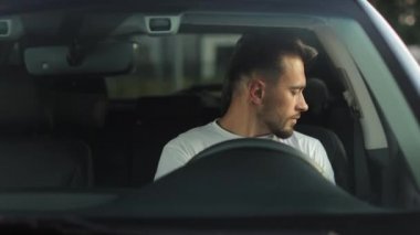 A man is sitting in the drivers seat of a car. He is closing the door and putting on his seat belt. He is putting his hands on the steering wheel. 4K 50fps