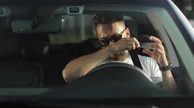 Gözlüklü bir adam bir arabanın sürücü koltuğunda oturuyor. Emniyet kemerini takıyor ve ellerini direksiyona koyuyor. O, kameraya bakıyor. 4K 50fps