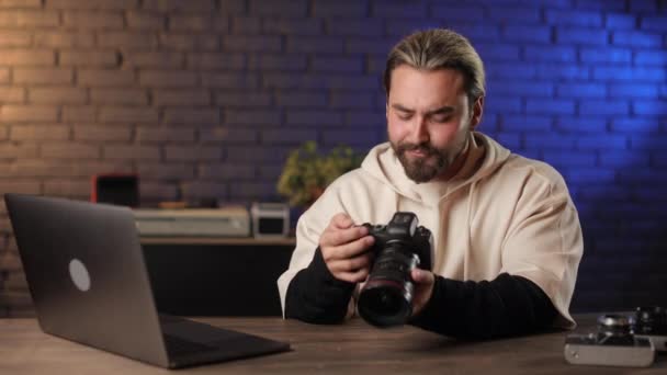 Mężczyzna siedzi przy biurku z laptopem i trzyma aparat fotograficzny — Wideo stockowe