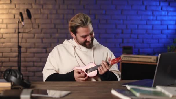 Šťastný muž sedí u stolu s notebookem a hraje ukulele — Stock video