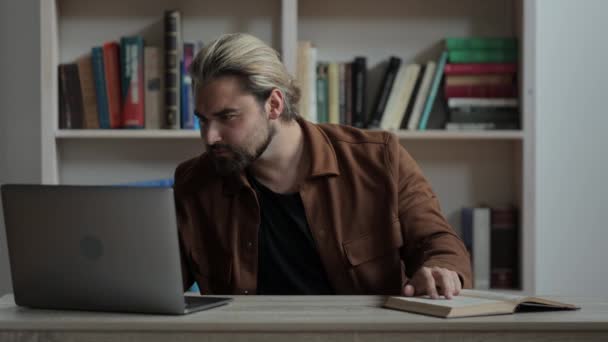 Handsome man reading book and typing on laptop — Stock Video