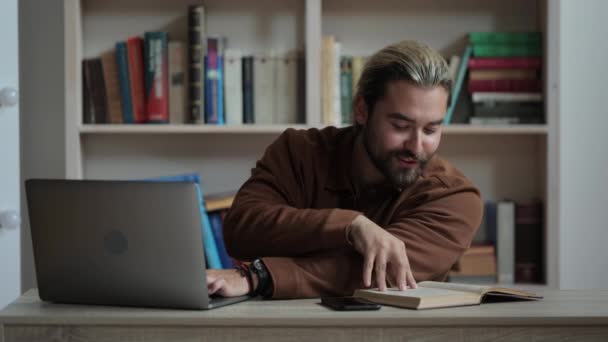 Homem positivo sentado à mesa com livro e usando laptop — Vídeo de Stock