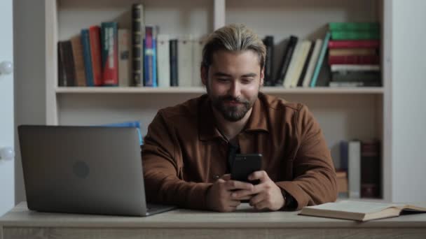 Hombre usando el móvil mientras está sentado en el escritorio con el ordenador portátil y libro — Vídeo de stock
