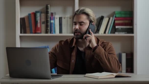 Homem barbudo falando no celular e digitando no laptop — Vídeo de Stock