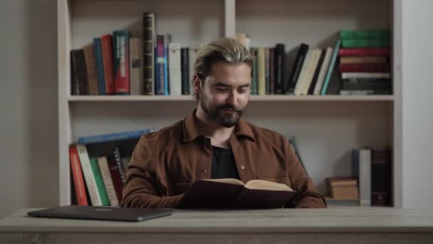 Blanke man aan het bureau met laptop en leesboek — Stockvideo