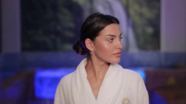 Beyaz, yumuşak bornozlu, sevimli genç bir kadının portresi gülümseyen ve modern otelin kaplıca alanındaki kameraya bakan. İnsan kavramı, tatil ve rahatlama.