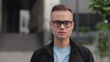 Gözlüklü genç bir adam başını kaldırıyor ve kameraya bakıyor. Gülümsüyor ve iş merkezinin yanında duruyor. 4k