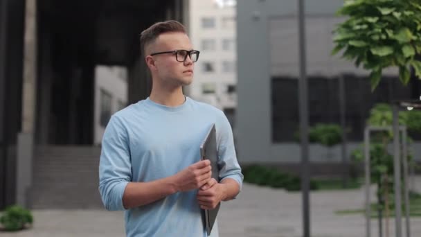 Een zakenman met een laptop in zijn handen staat en kijkt weg. Hij draagt een bril en een trui. Hij staat in de buurt van het zakencentrum. 4k — Stockvideo