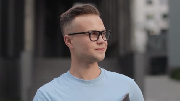 Un hombre de negocios con un portátil en sus manos está de pie y mirando a la cámara. Lleva gafas y un suéter. Él sonríe. Está parado cerca del centro de negocios. Disparos de cerca. 4k — Vídeos de Stock