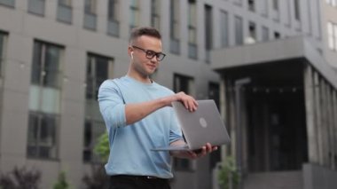 Genç bir adam iş merkezinin yanında duruyor. Bilgisayarını kapatıyor ve kameraya bakıyor. Gülümsüyor. O gözlük ve bir kazak giyiyor. 4k