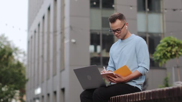 Młody mężczyzna siedzi na ławce w pobliżu centrum biznesowego. Pracuje nad laptopem i robi notatki w notatniku. Nosi okulary. 4k — Wideo stockowe