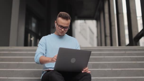 Een jongeman zit op een zakencentrum. Hij werkt aan een laptop en maakt aantekeningen in een notitieboekje. Hij draagt een bril. Hij steekt zijn hoofd op en kijkt weg. 4k — Stockvideo