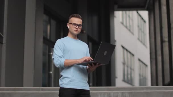 Een jongeman staat op een trap. Hij werkt aan een laptop. Hij steekt zijn hoofd op en kijkt naar de camera. Hij glimlacht. Hij draagt een bril en een trui. 4k — Stockvideo