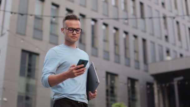 Een jonge zakenman komt in de buurt van het zakencentrum. Hij houdt een laptop in zijn handen en sms 't op zijn smartphone. 4k — Stockvideo