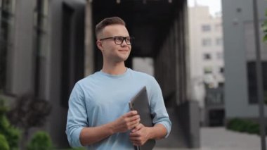 Elinde dizüstü bilgisayarı olan bir iş adamı ayakta kameraya bakıyor. O gözlük ve bir kazak giyiyor. Gülümsüyor. İş merkezinin yanında duruyor. 4k