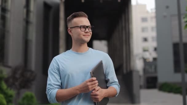 Een zakenman met een laptop in zijn handen staat naar de camera te kijken. Hij draagt een bril en een trui. Hij glimlacht. Hij staat in de buurt van het zakencentrum. 4k — Stockvideo