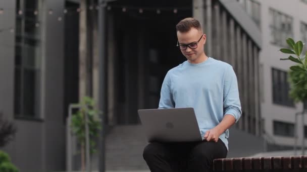 Een jongeman zit op een bankje in de buurt van het zakencentrum. Hij werkt aan een laptop en lacht. Hij steekt zijn hoofd op en kijkt weg. Hij draagt een bril. 4k — Stockvideo