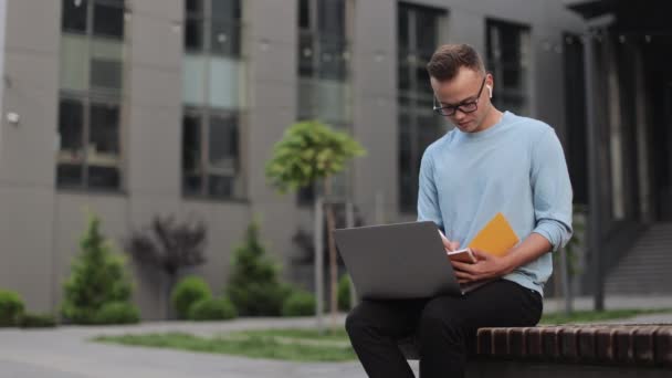 Um jovem está sentado num banco perto do centro de negócios. Ele está trabalhando em um laptop e fazendo anotações em um notebook. Ele está usando óculos. Ele está levantando a cabeça e desviando o olhar. 4k — Vídeo de Stock