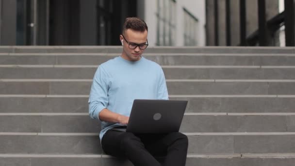 Een jongeman zit op een zakencentrum. Hij werkt aan een laptop. Hij draagt een bril en een trui. De camera draait om hem heen. 4k — Stockvideo