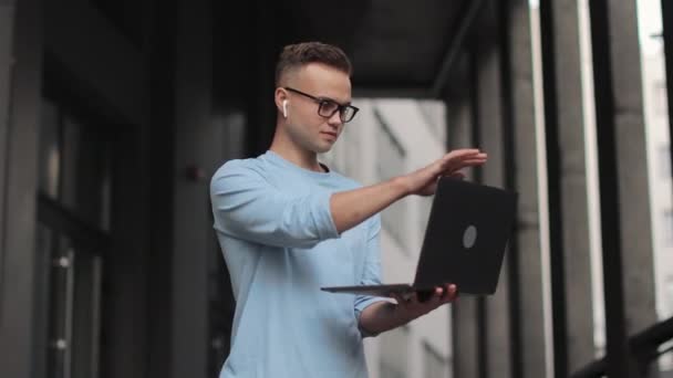 Młody człowiek stoi na schodach w centrum biznesowym. Zamyka laptopa i patrzy w kamerę. Uśmiecha się. Nosi okulary i sweter. 4k — Wideo stockowe
