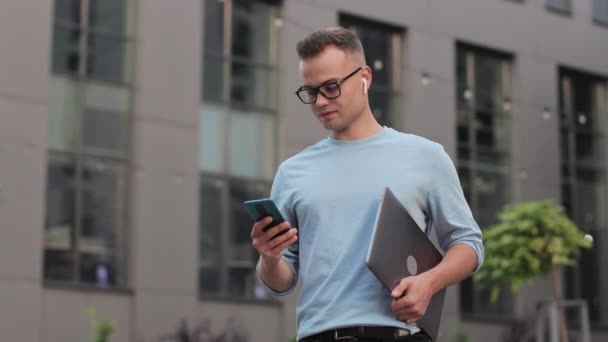 Молодой бизнесмен стоит рядом с бизнес-центром. Он держит в руках ноутбук и пишет смс на смартфоне. Он улыбается и поднимает голову. 4k — стоковое видео