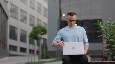 Genç bir adam iş merkezinin yanındaki bankta oturuyor. Bilgisayarı açıyor ve çalışmaya başladı. O gözlük takıyor. 4k