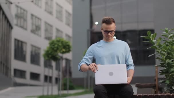 한 젊은이가 사업 중심지 근처에 있는 벤치에 앉아 있다. 그는 노트북을 열고 작동하기 시작 했습니다. 안경을 쓰고 있다. 4k — 비디오