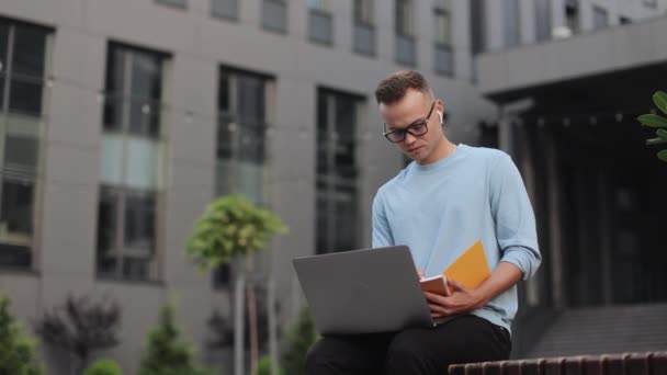 Młody mężczyzna siedzi na ławce w pobliżu centrum biznesowego. Pracuje nad laptopem i robi notatki w notatniku. Nosi okulary. Kamera porusza się od prawej do lewej wokół niego. 4k — Wideo stockowe