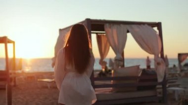 Yaz günbatımında kumlu sahilde yürüyen genç bir kadın.