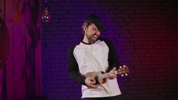 Joyful man actively dancing in studio with ukulele in hands — Stock video