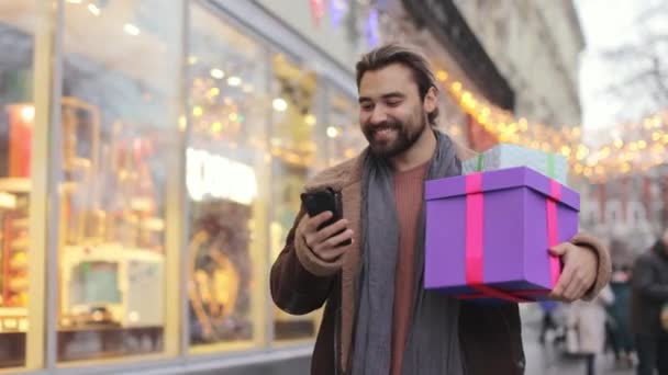 Man with mobile and christmas gifts walking on city street — Stockvideo